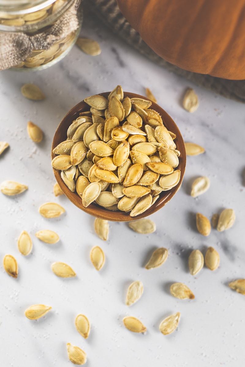 An overflowing bowl of pumpkin seeds with an orange pumpkin and a jar of more seeds.