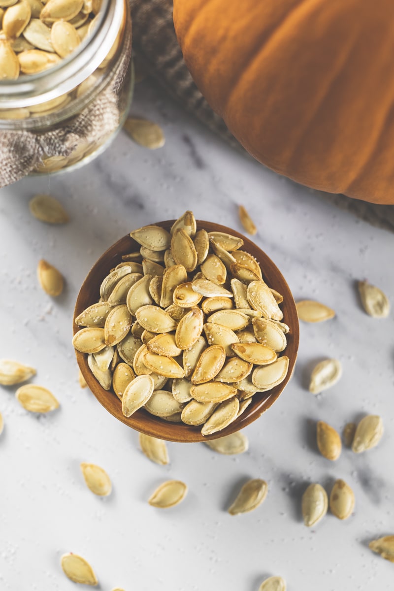 An overflowing bowl of roasted pumpkin seeds beside a pumpkin and mason jar full of more seeds.