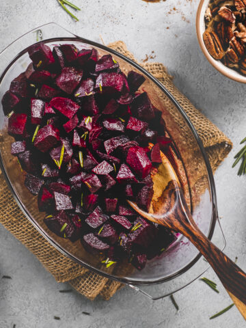 A wooden serving spoon resting in a roasting dish full of baked balsamic glazed beets topped with fresh rosemary.