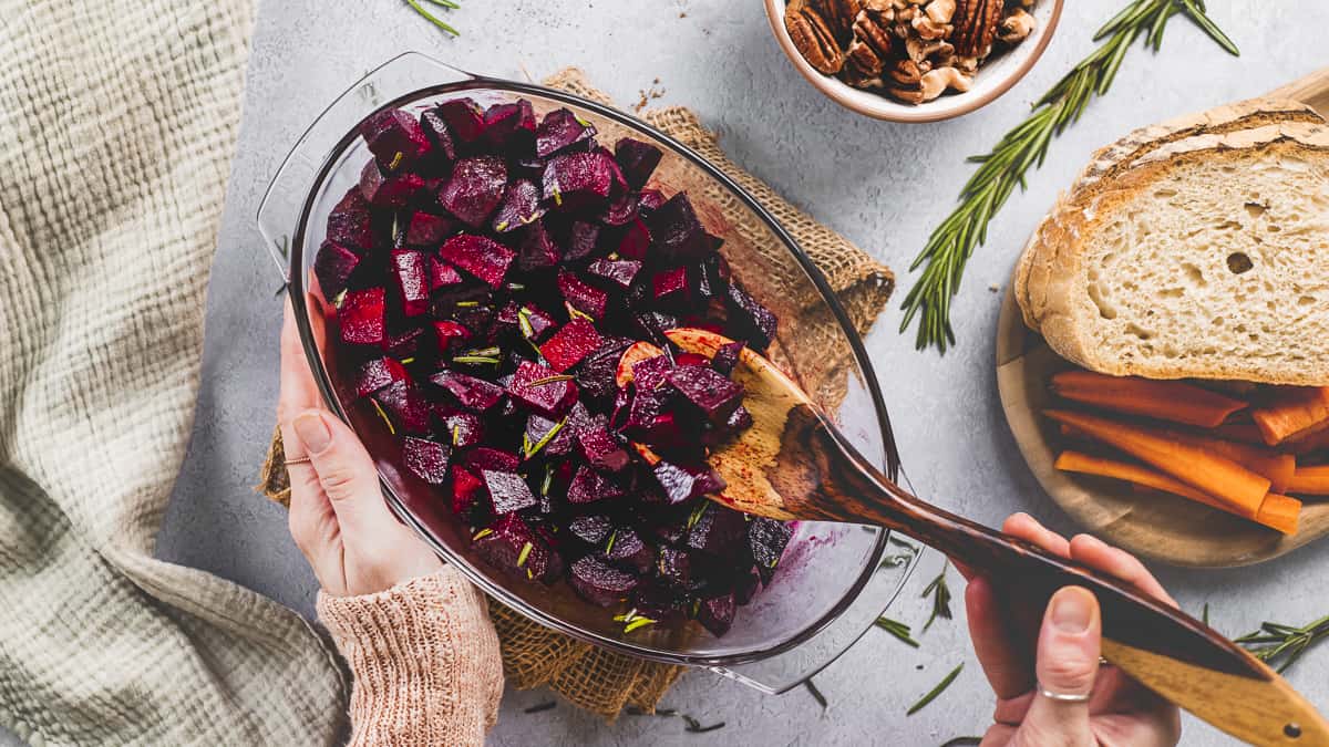 Roasted beets being scooped out of a baking dish beside a sprig of rosemary, a bowl of nuts and a platter of bread and vegetables.