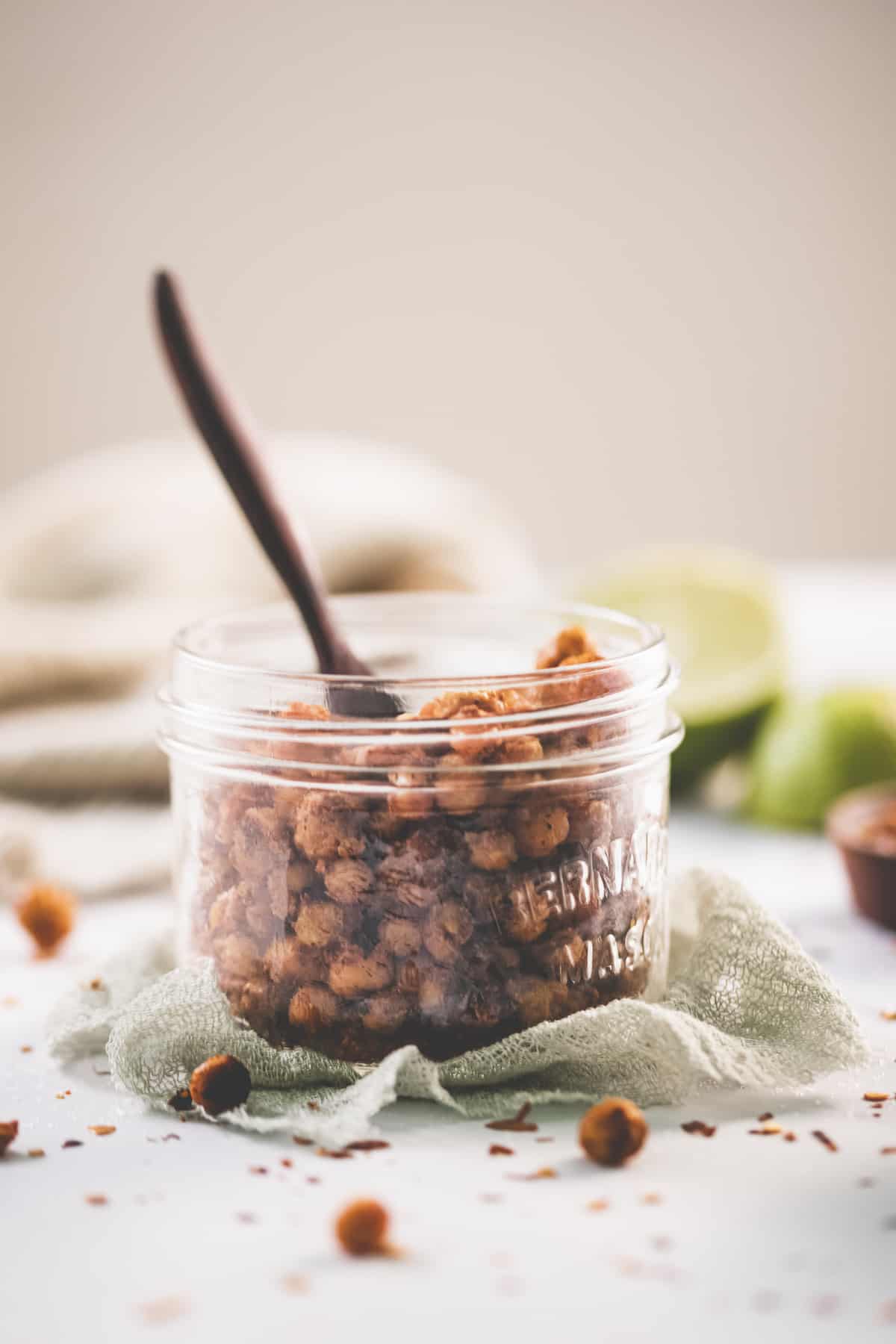 Chili Lime Chickpeas in a glass jar with a wooden serving spoon, surrounded by a bowl of chili flakes, fresh lime and spilt crumbs.