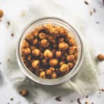 A jar of generously seasoned chili lime chickpeas surrounded by chili flake crumbs and spilt chickpeas.