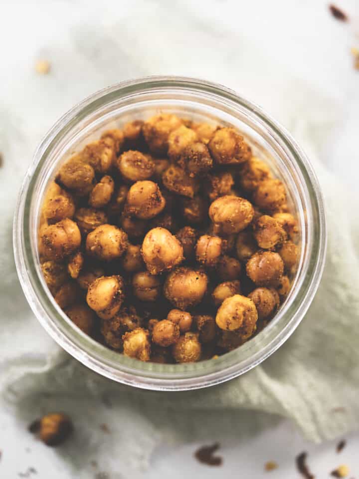 A jar of generously seasoned chili lime chickpeas surrounded by chili flake crumbs and spilt chickpeas.