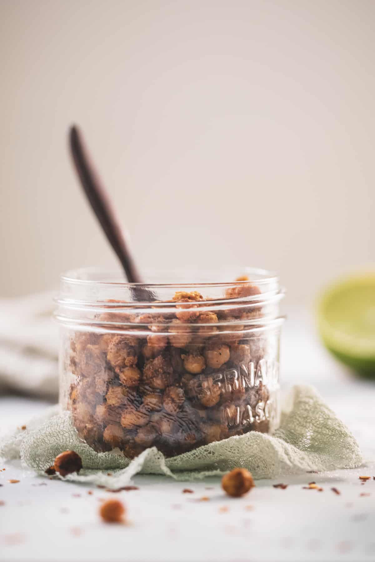 A jar of roasted chickpeas served on a sage green cloth with a wooden serving spoon, fresh lime and spilt chickpeas surrounding.