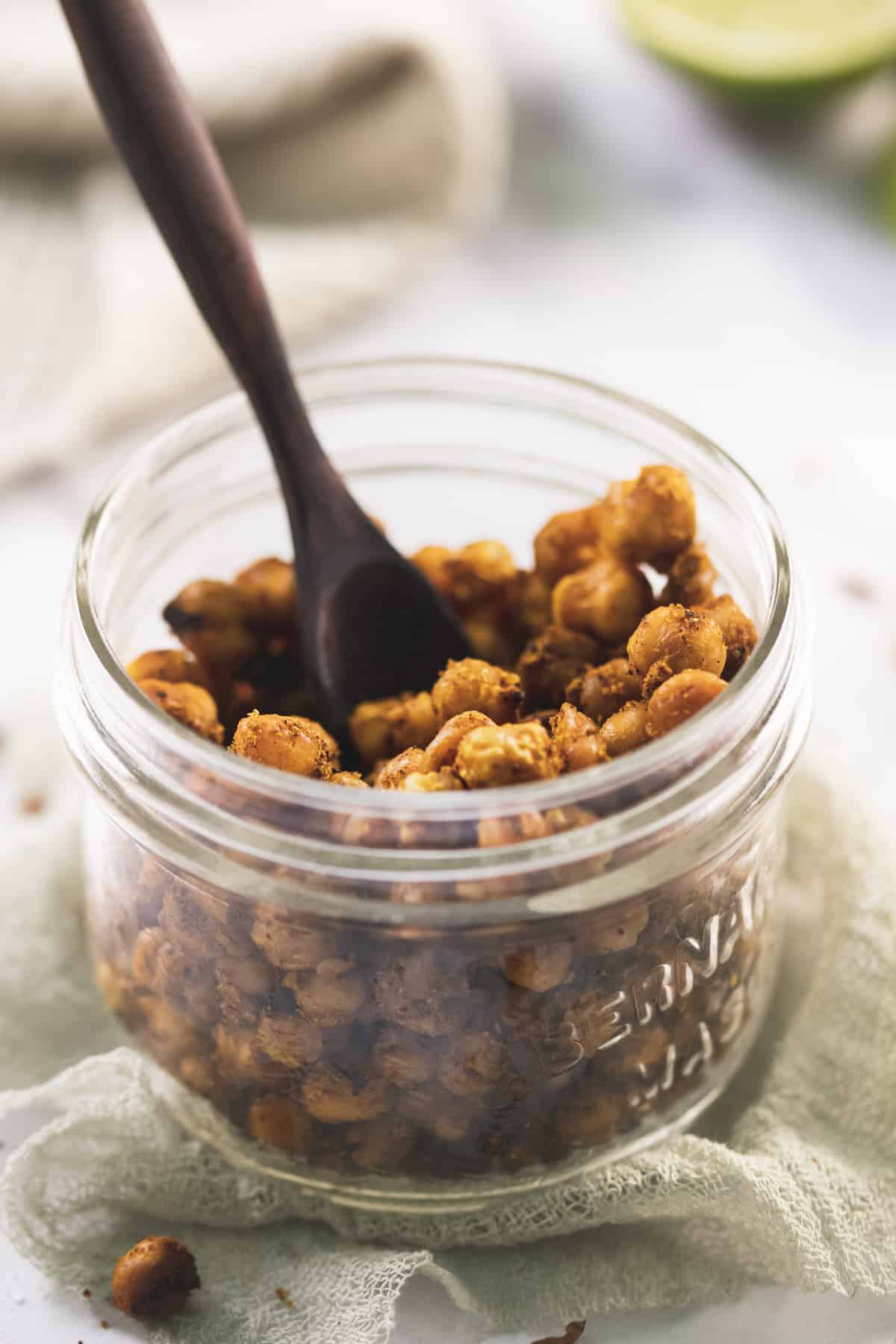 A wooden spoon resting in a jar for roasted chickpeas on a sage cheesecloth with spilt crumbs surrounding and a freshly sliced lime in the background.