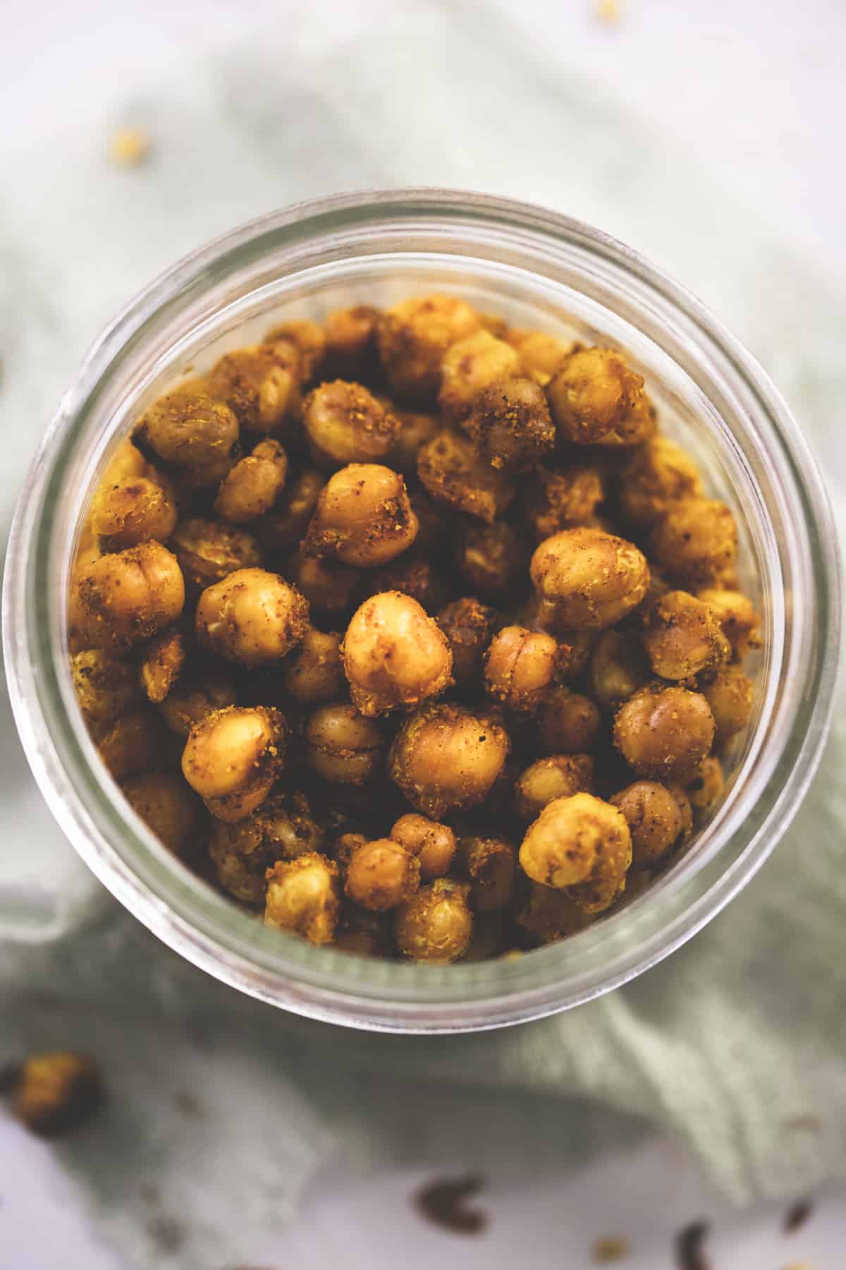 Roasted and seasoned chickpeas in a glass jar resting on a sage cheesecloth with split chickpeas and chili flakes surrounding.