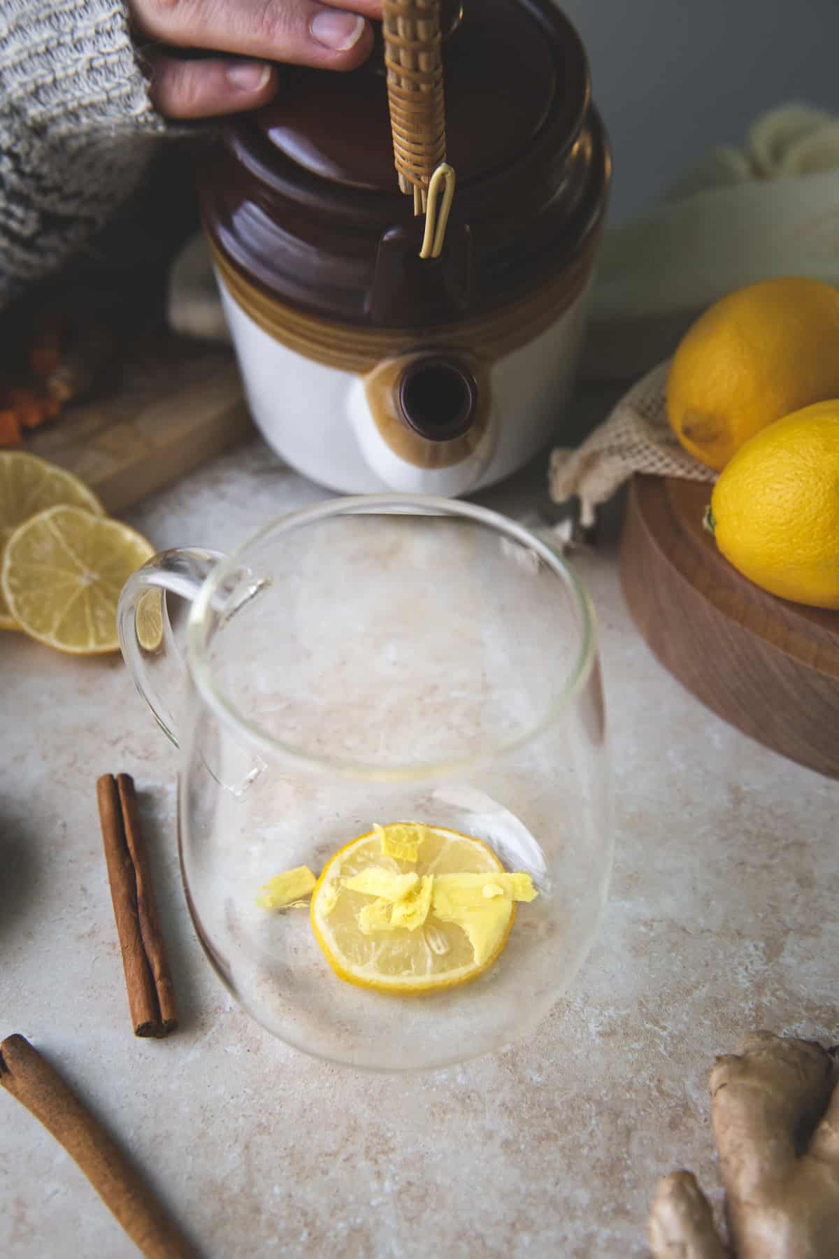 A teapot being lifted to pour hot water over a fresh lemon round and sliced ginger root that are resting in the bottom of a glass mug.