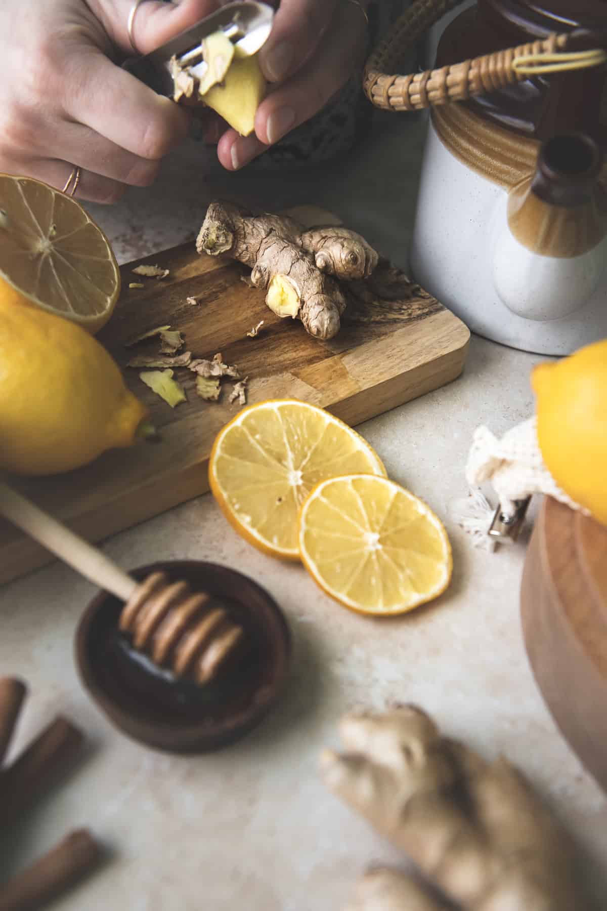 Ginger root being peeled over a cutting board with sliced and whole lemon, a ceramic teapot and a honey dipper resting in a small wooden bowl.