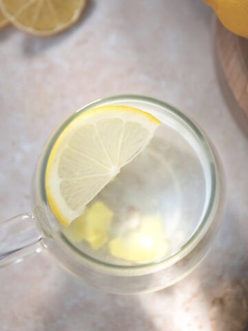A half slice of lemon floating in a mug of water with chopped ginger root settled at the bottom. Sitting beside whole and sliced lemon resting on a cutting board.