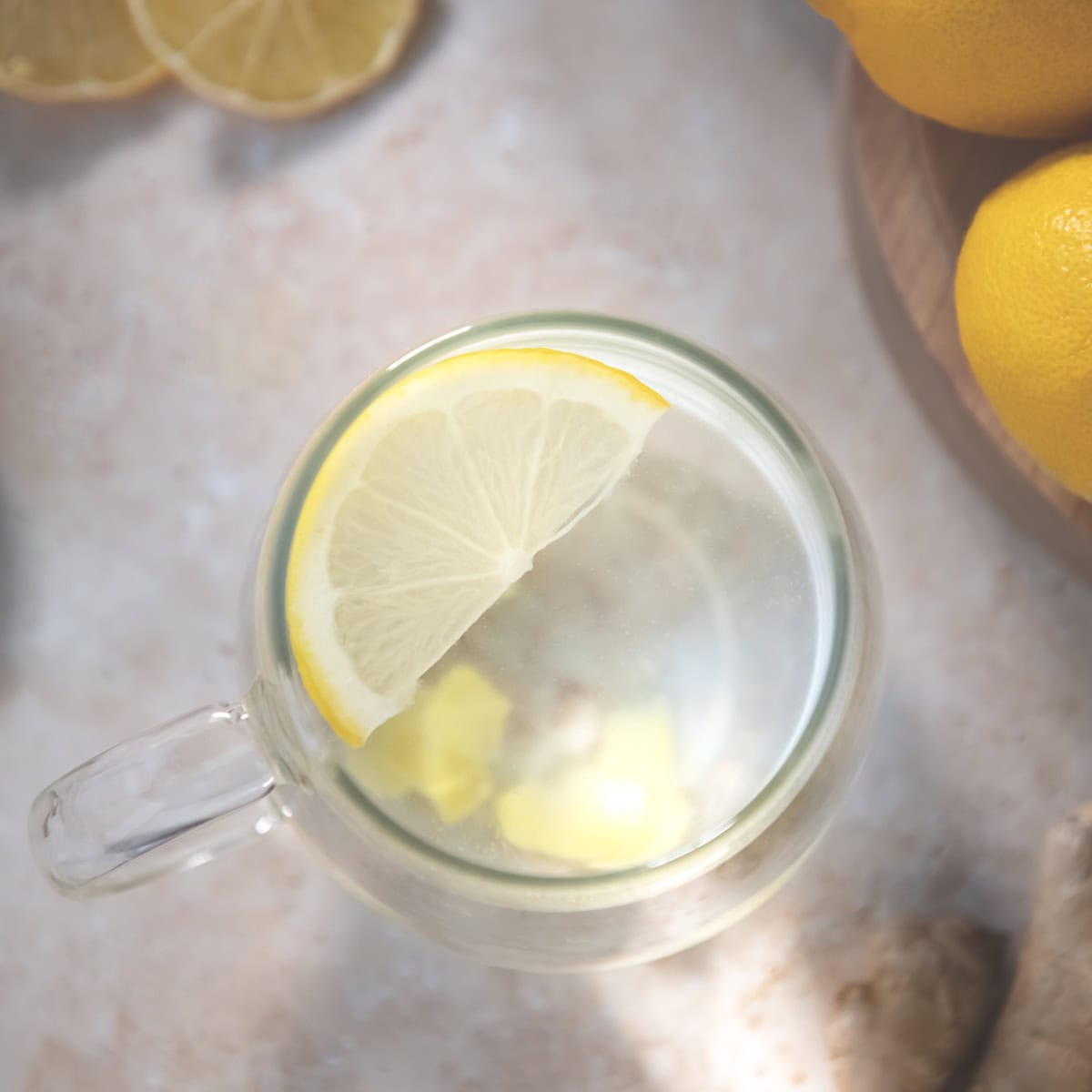 A half slice of lemon floating in a mug of water with chopped ginger root settled at the bottom. Sitting beside whole and sliced lemon resting on a cutting board.