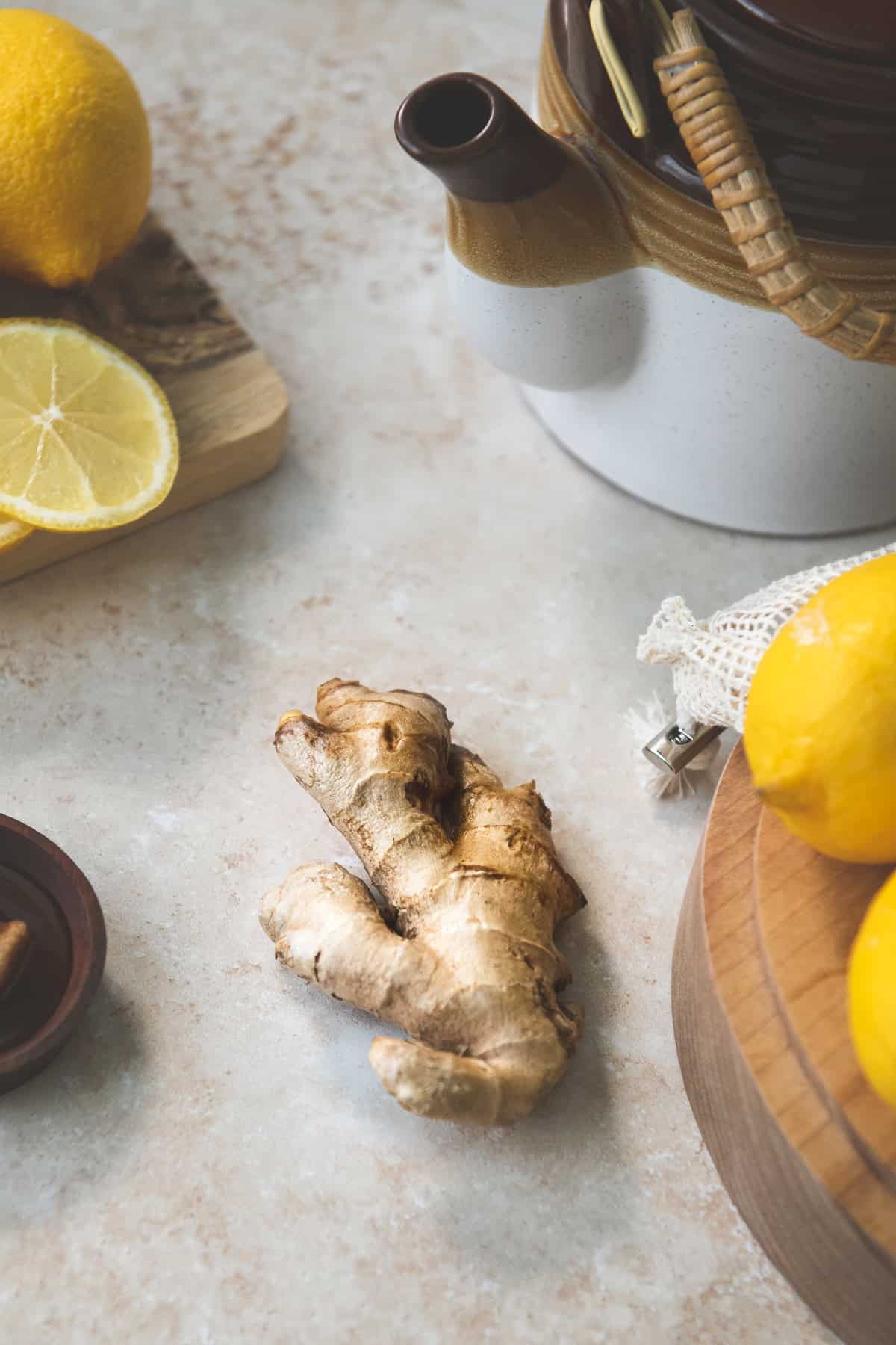 A hunk of fresh ginger root centred between a spoon holder, ceramic teapot, and sliced lemon resting on a cutting board.