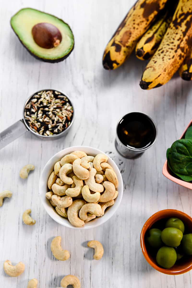 An overflowing bowl of raw cashews surrounded by other foods, including whole grain wild rice, olives, olive oil, bananas and avocado.