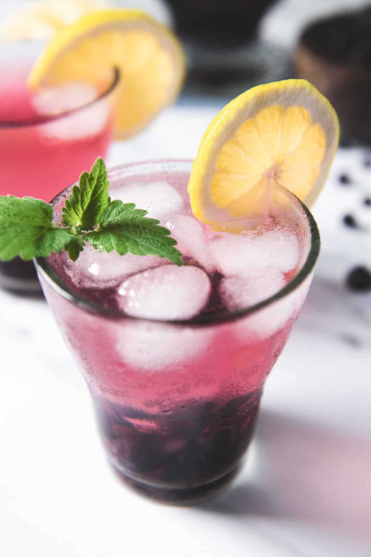 Two glasses of blueberry lemon-infused water with ice, lemon slices, and a sprig of lemon balm.