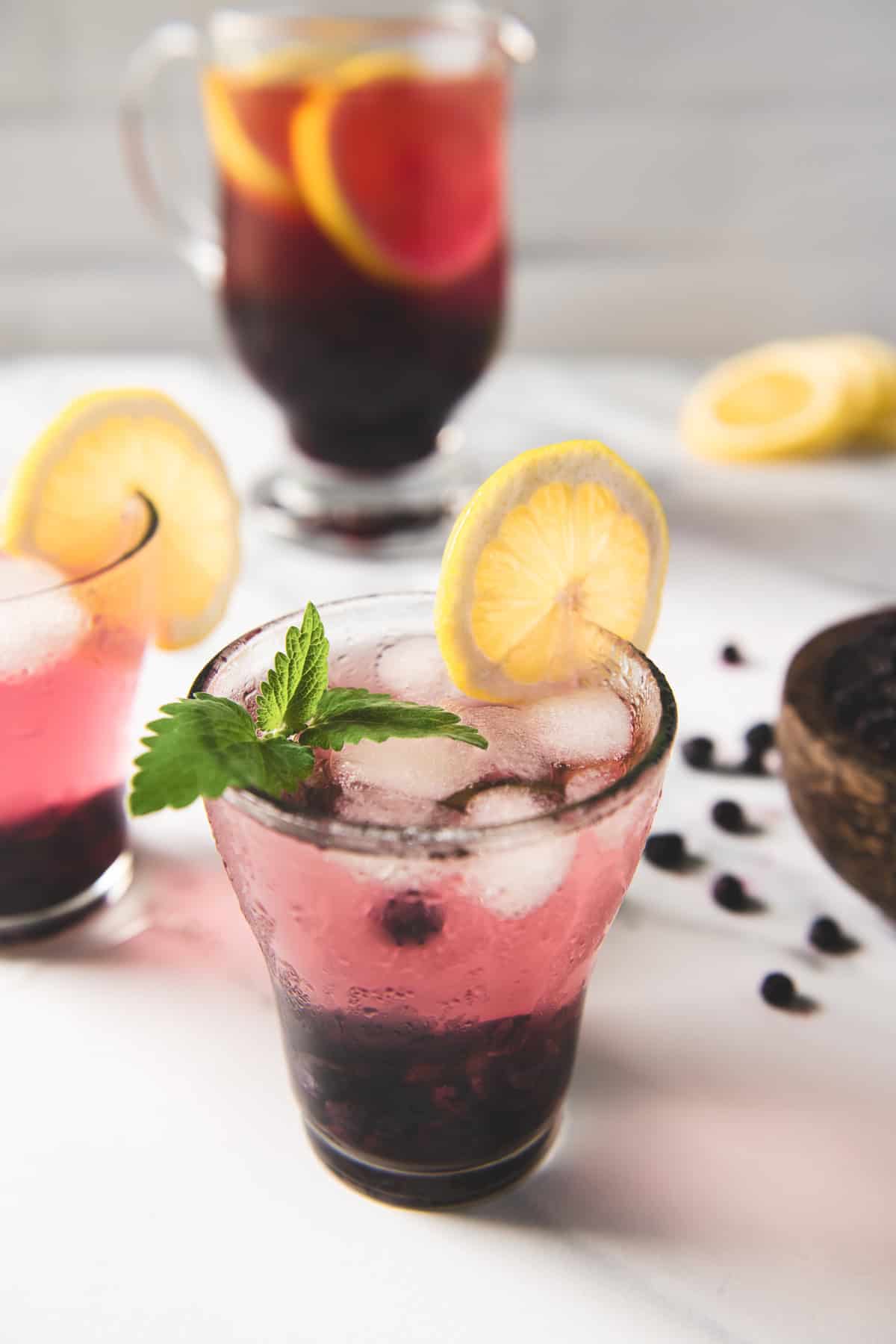 A pitcher of infused blueberry lemon water beside two glasses garnished with lemon slices and lemon balm.