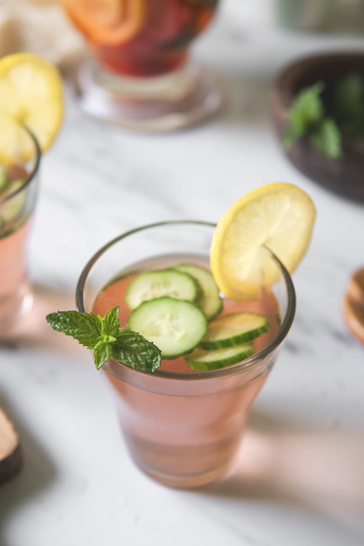 A glass of pink strawberry water topped with cucumber slices and garnished with a lemon round and a sprig of mint.