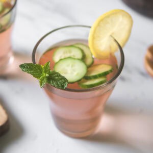 A glass of pink strawberry-infused water garnished with sliced cucumber, lemon, and a sprig of mint.