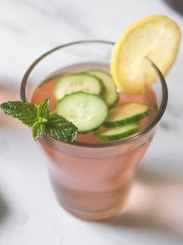 A glass of pink strawberry-infused water garnished with sliced cucumber, lemon, and a sprig of mint.