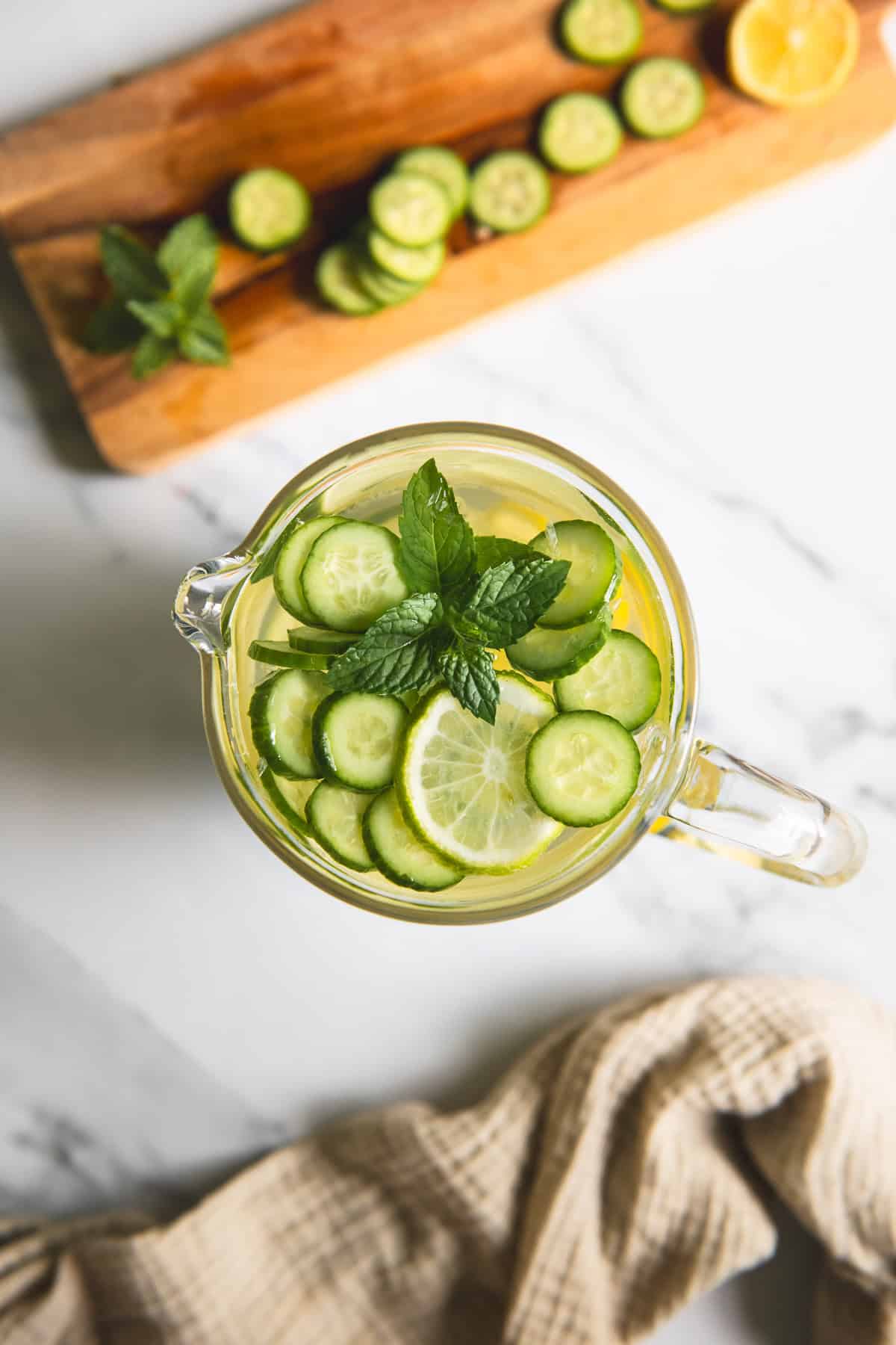 Sliced cucumber and lime floating with a sprig of mint in a pitcher of water.