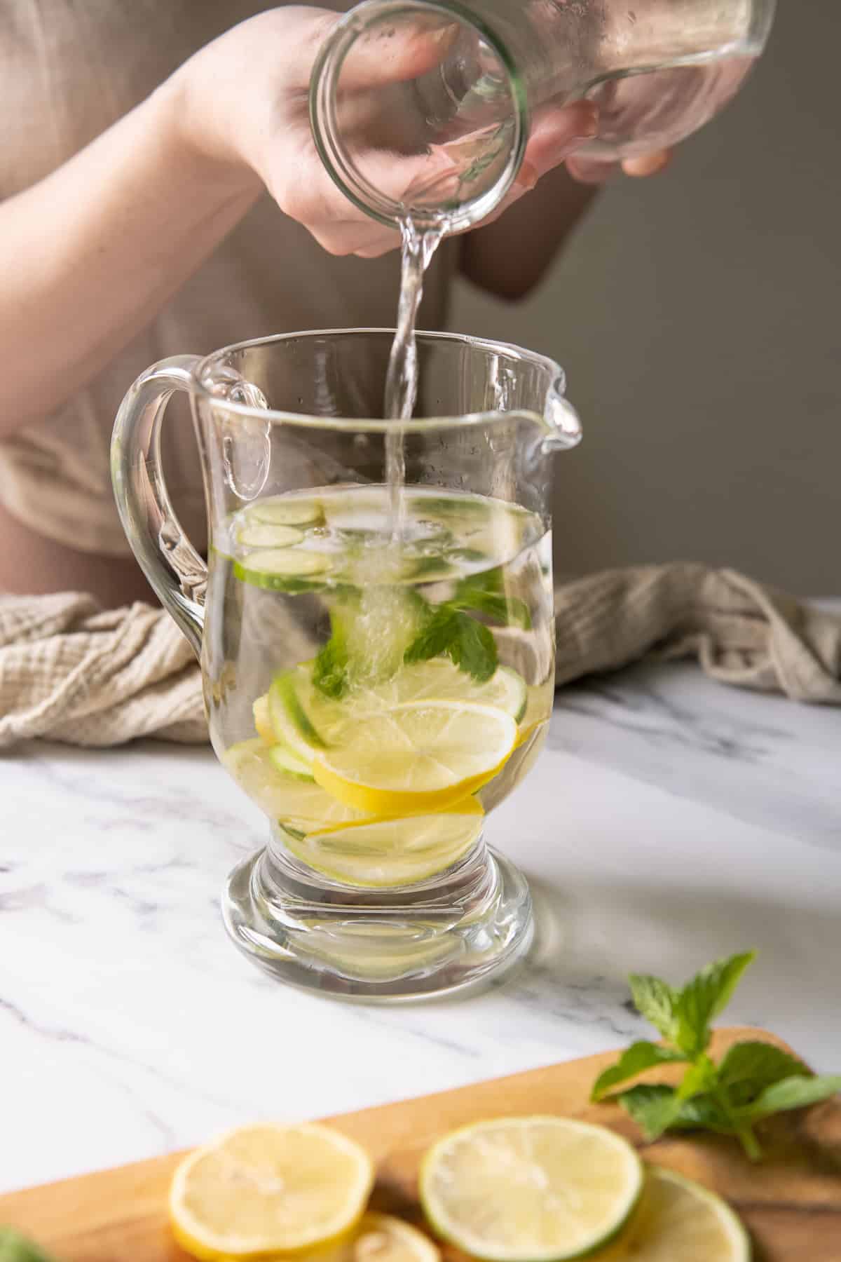 A woman pouring water over sliced cucumber, lemon, lime and fresh mint.