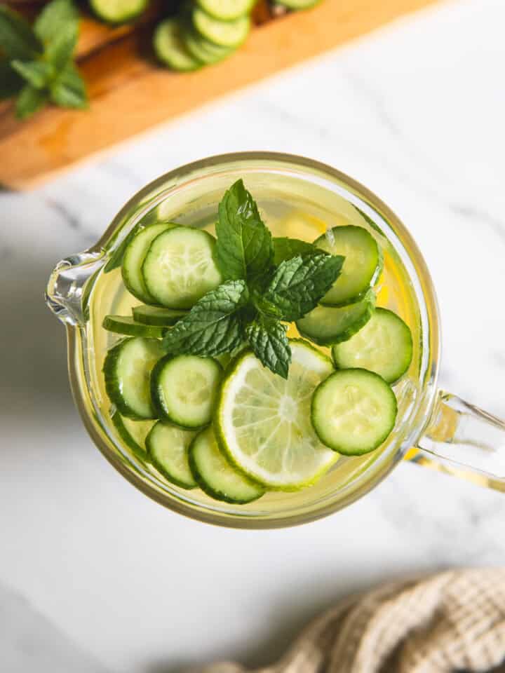 Slices of cucumber and lime floating in a pitcher of water with a sprig of mint.