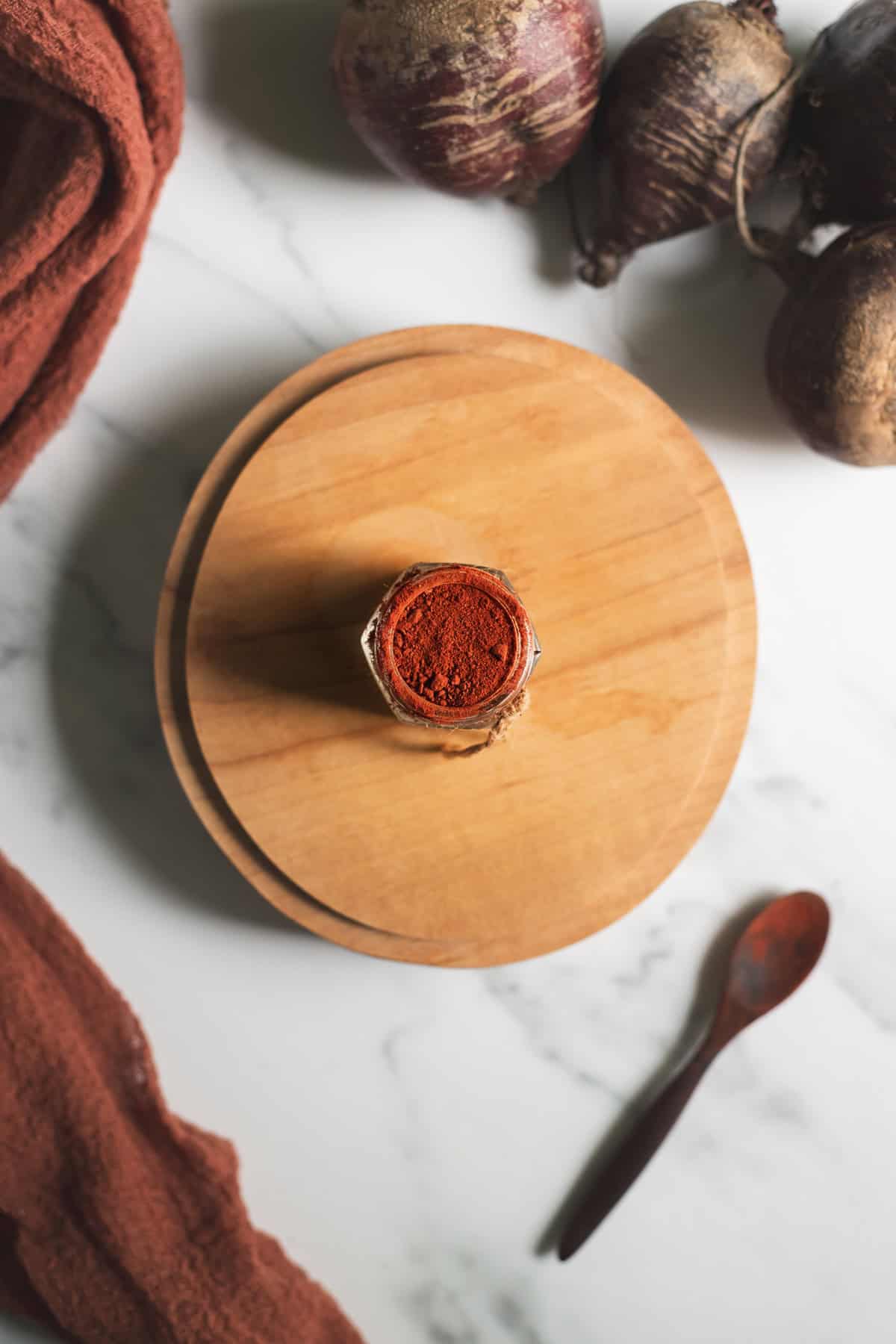 A jar of beetroot powder surrounded by fresh beets, a wooden teaspoon and a linen cloth.