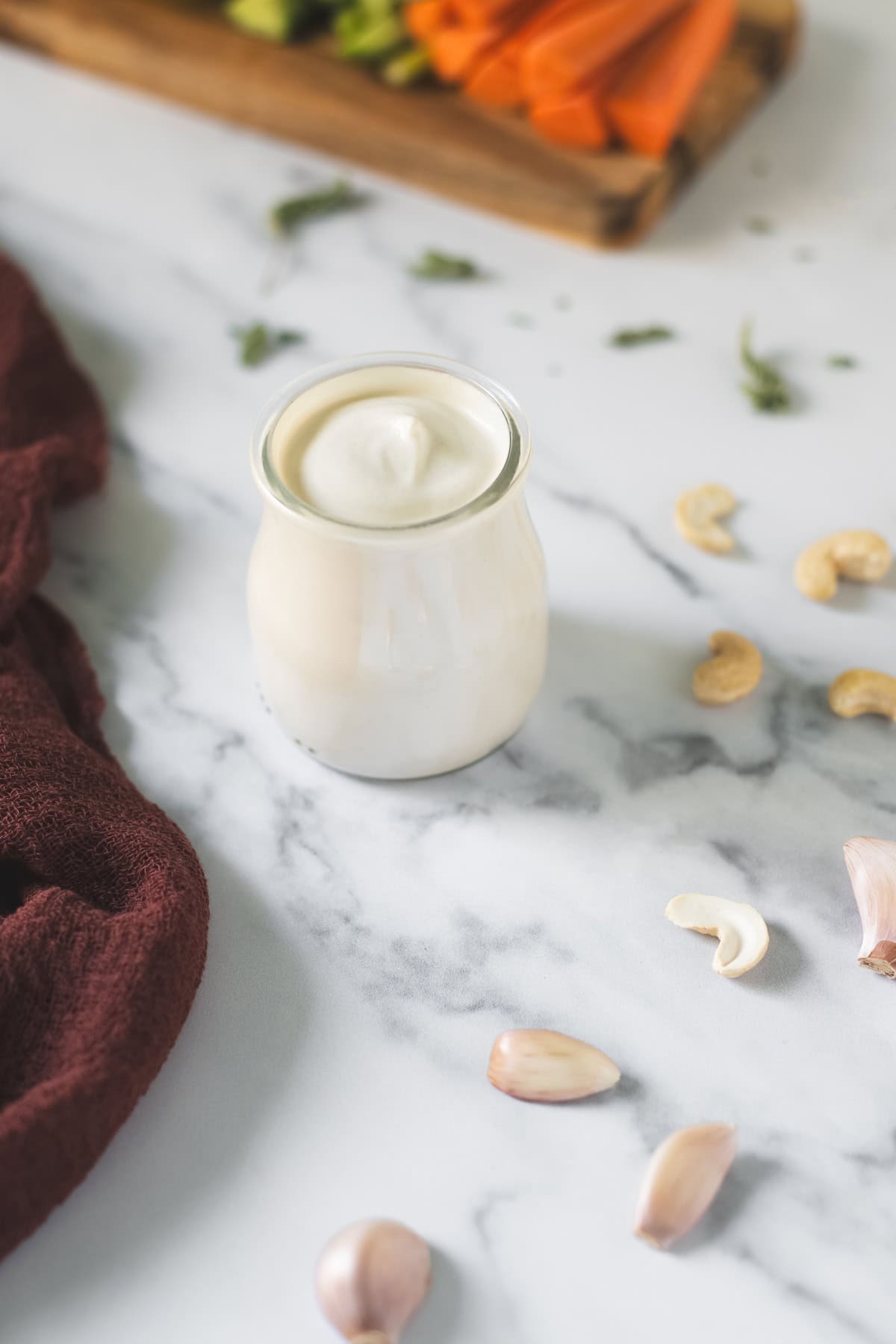 A jar of creamy cashew dip surrounded by raw cashews, garlic cloves and a tray of carrot and cucumber sticks.