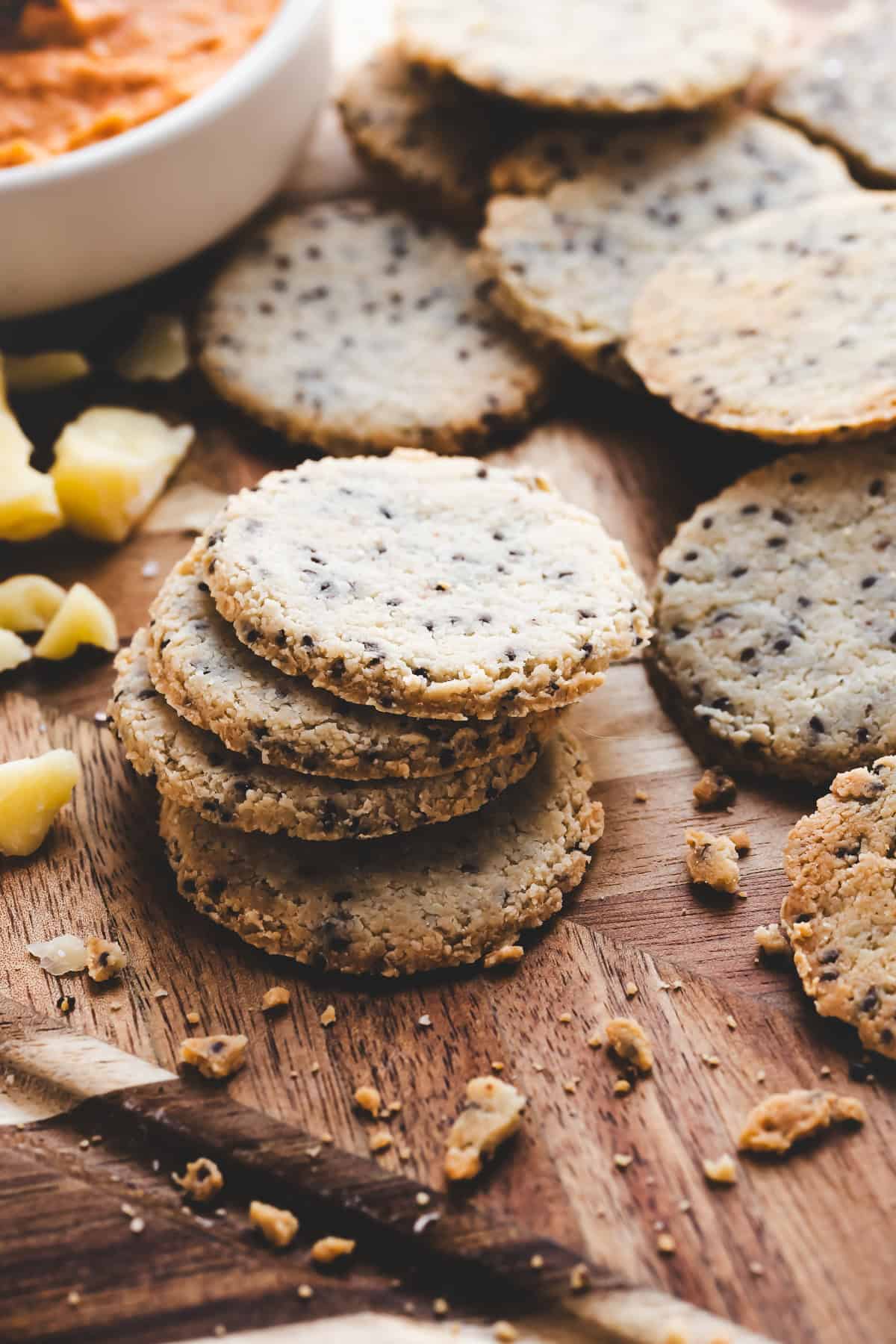 Chia seed crackers stacked on a serving board beside cheese and a bowl of dip.