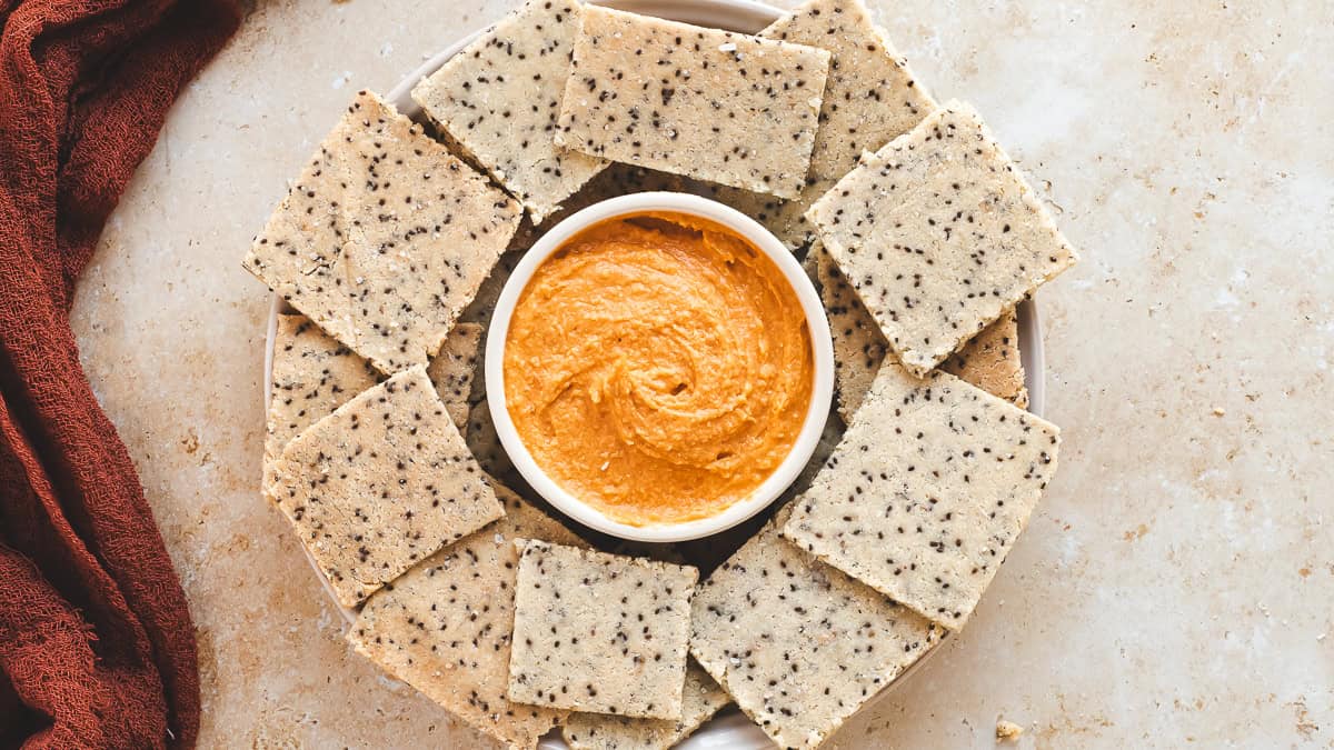 A plate of chia seed crackers with red pepper hummus in the centre.
