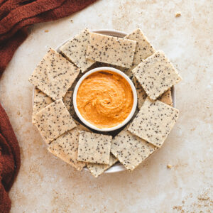 A platter of chia seed crackers with a bowl of hummus in the middle.