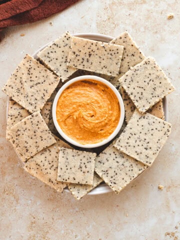 A platter of chia seed crackers with a bowl of hummus in the middle.