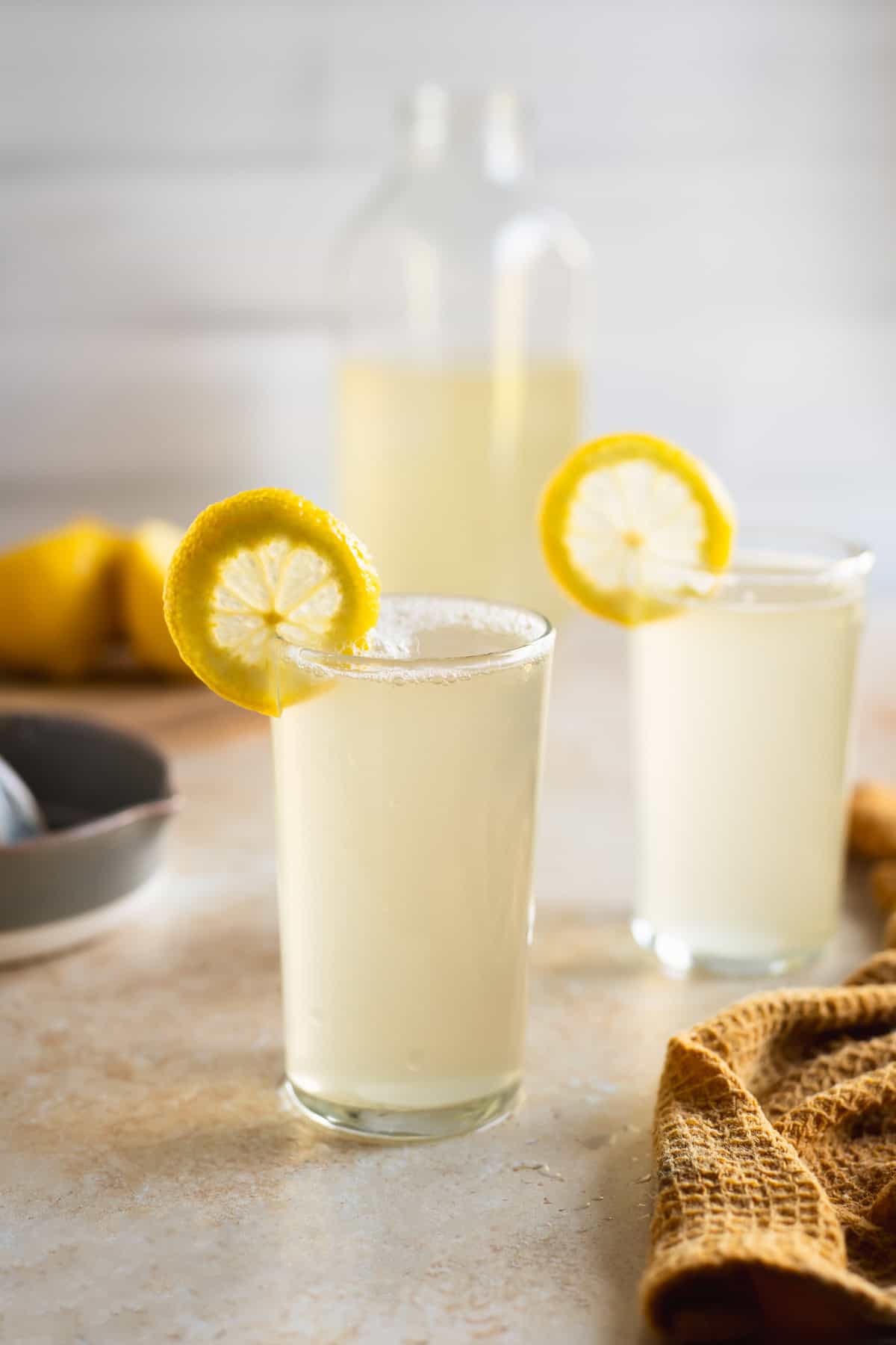 A partially full jug of lemon soda behind two full glasses garnished with lemon rounds.