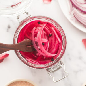 An open jar of pink, jewel-toned pickled red onions with whole peppercorns and a serving fork.