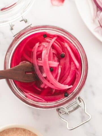 An open jar of pink, jewel-toned pickled red onions with whole peppercorns and a serving fork.