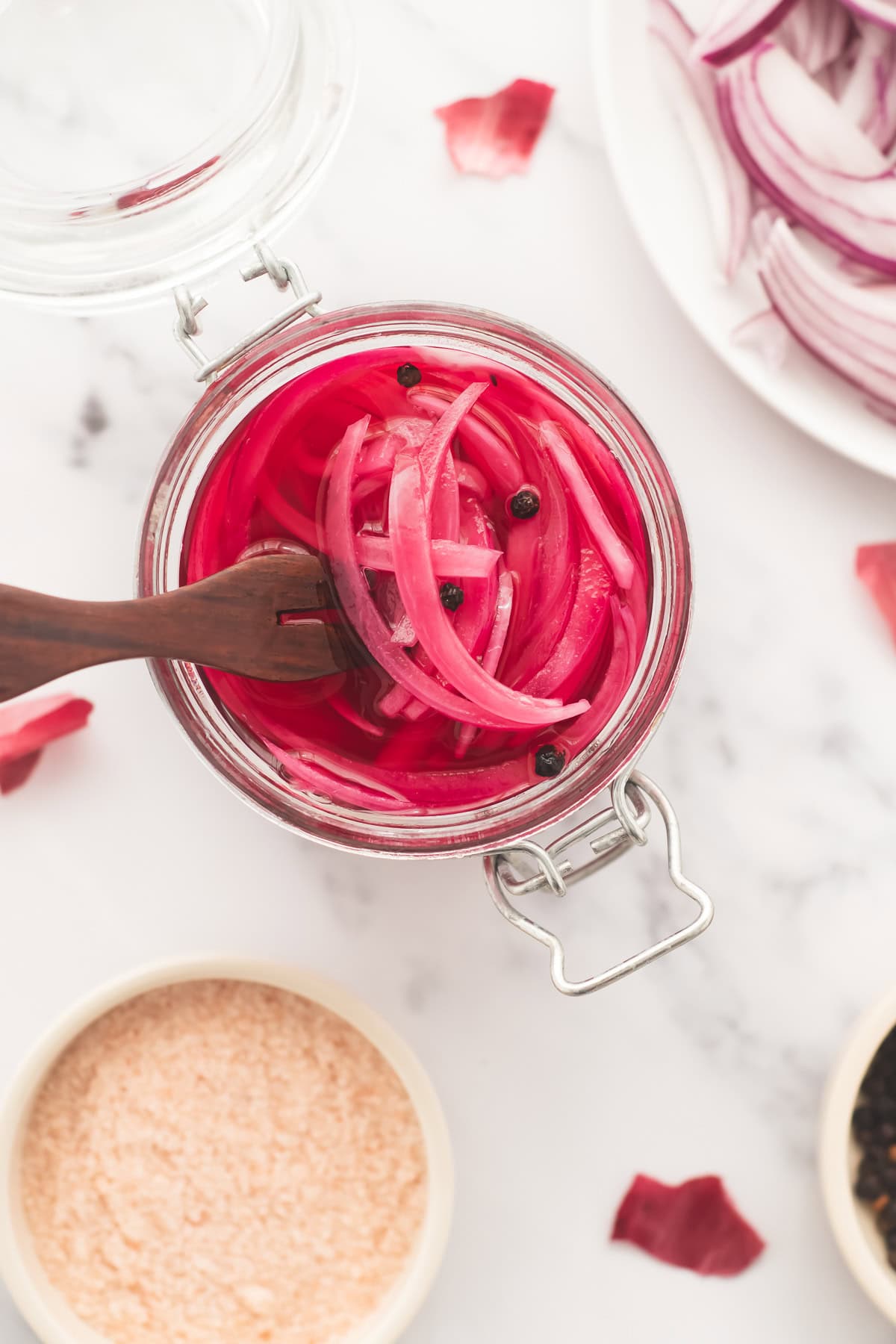 An open jar of pink, jewel-toned pickled red onions with whole peppercorns and a serving fork.