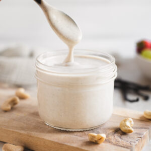 A glass jar of vanilla-speckled cashew crème and a wooden spoon lifted from the jar, showcasing a smooth drip of cream falling back into the jar.