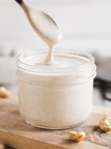 A glass jar of vanilla-speckled cashew crème and a wooden spoon lifted from the jar, showcasing a smooth drip of cream falling back into the jar.