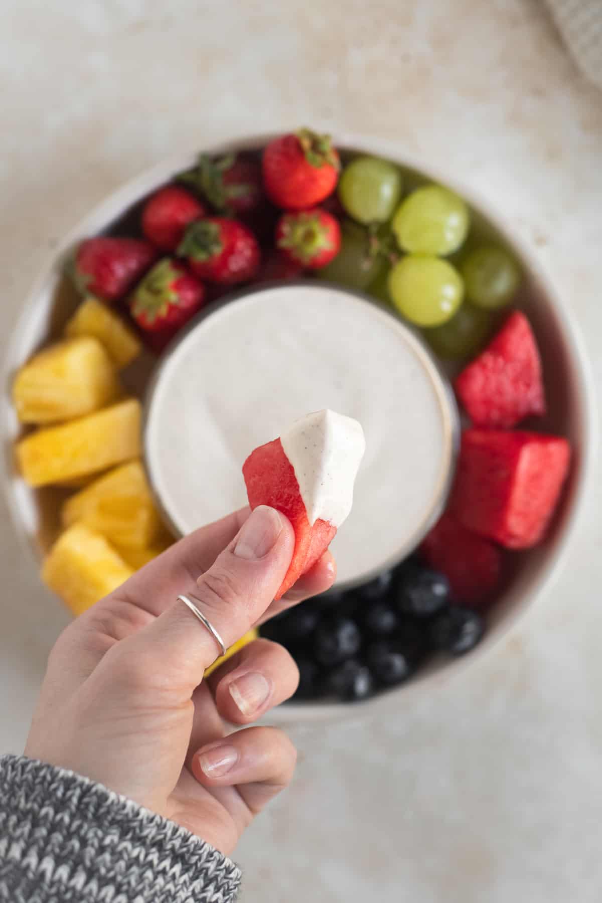 A vibrant slice of pink watermelon with creamy dairy-free sweet crème coating one corner, held up in a hand over a round platter of fresh fruit with a bowl of dip in the centre.