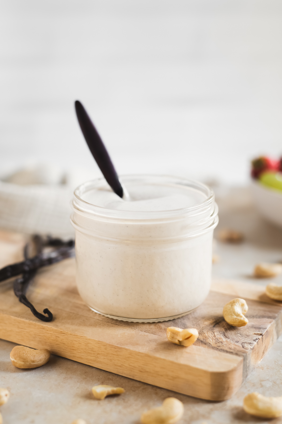 A glass jar filled with off-white, vanilla-speckled cashew creme surrounded by raw cashews, vanilla beans and a bowl of fruit.