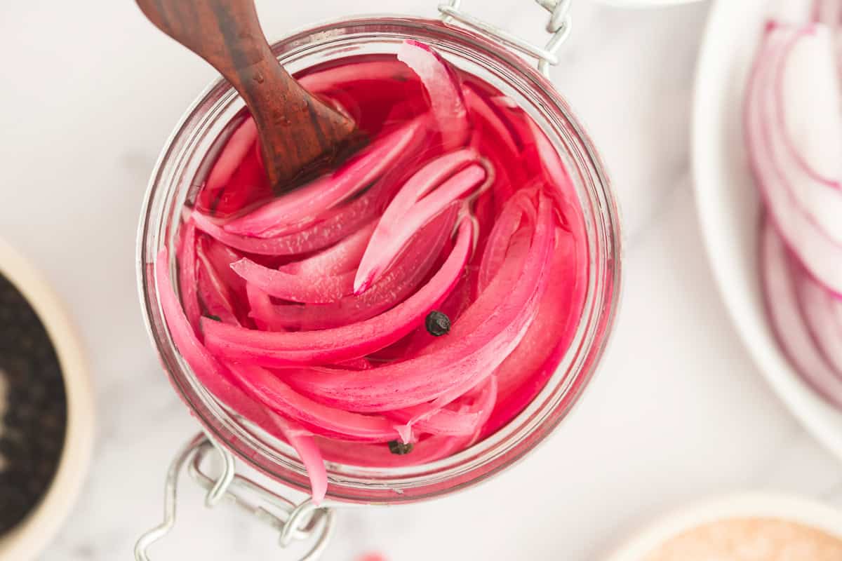 A wood tasting fork submerged in a jar of pink pickled red onion.